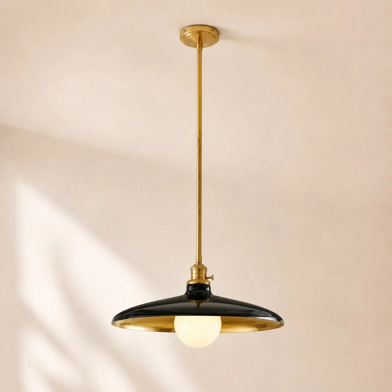 A modern pendant light fixture with a black dome-shaped shade, gold-toned metal frame and canopy, and a glowing warm white globe bulb, suspended from a ceiling against a warm beige wall.