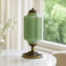 Decorative green vase with gold base on a table near a window with white curtains.