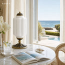 Decorative lamp on a table with an open book, glass of water, and flowers in a room with a view of the ocean.