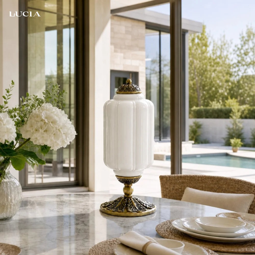 Decorative white lamp with gold base on a table in a modern indoor setting with large windows.