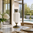 Decorative white lamp with gold base on a table in a modern indoor setting with large windows.