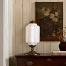 Decorative table lamp with white glass shade and gold base on a wooden surface, with a painting of flowers in the background.