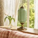 Decorative green lamp with gold base on a wooden surface near a window with white curtains.