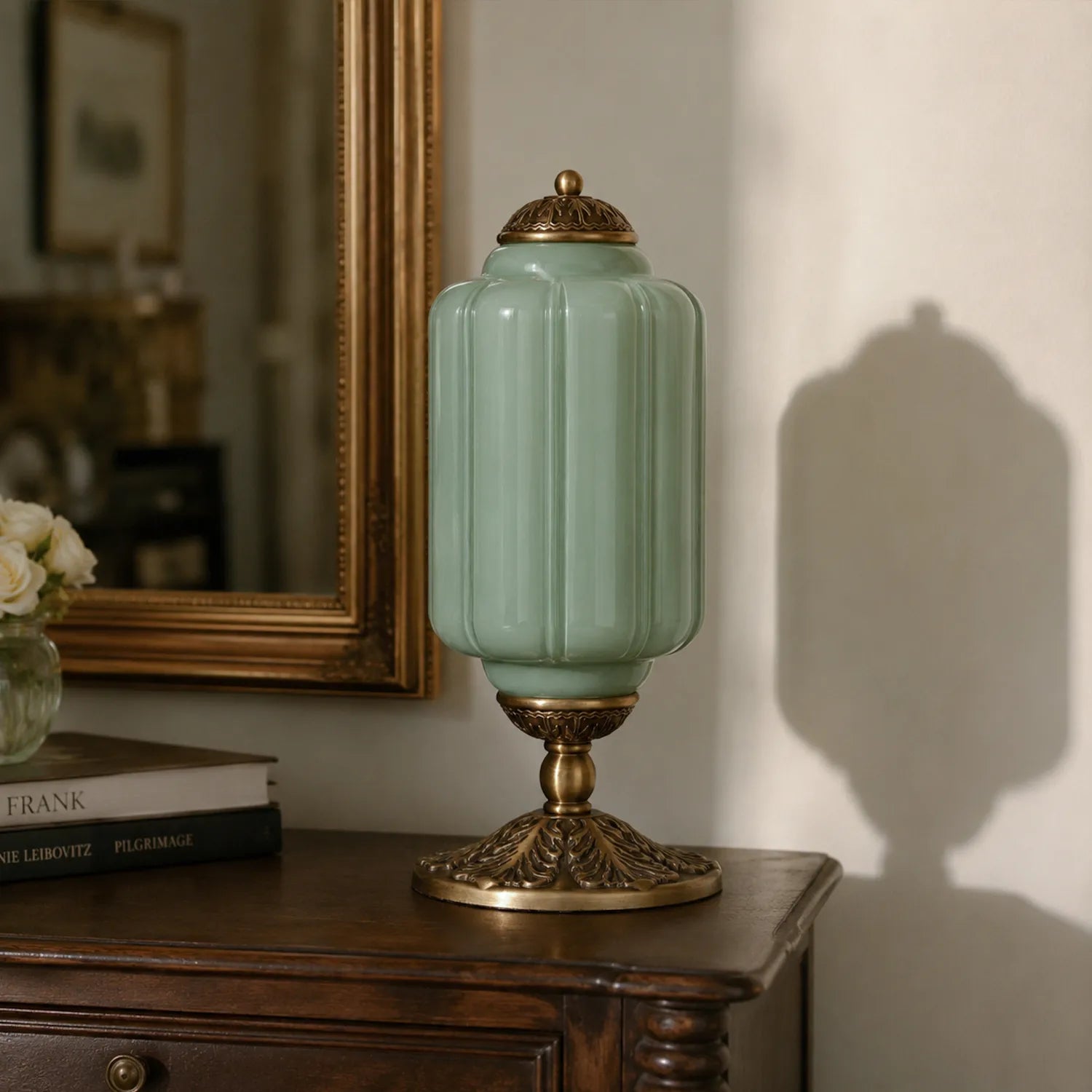 Decorative green lamp with gold base on a wooden surface, with books and a mirror in the background.