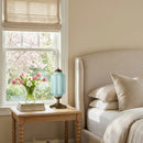 Neatly made bed with pillows next to a window with a view of trees, a wooden side table with books and a vase of flowers.