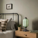 Bedroom with a wooden nightstand featuring a green lamp and books, next to a bed with pillows.