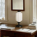 Table lamp on a desk with a laptop and books in a room with a window and mirror.