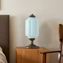 Table lamp with a blue glass shade on a wooden surface next to a chair and headboard.