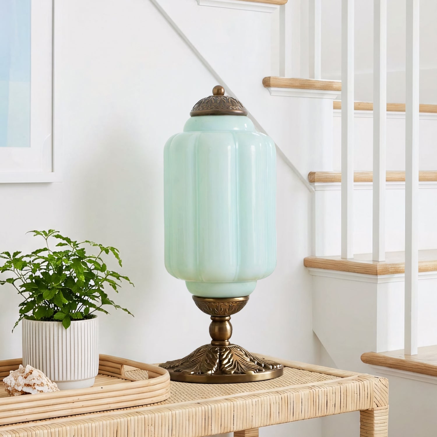 Mint green table lamp with brass base on a wooden side table next to a plant, with a staircase in the background.
