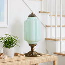 Mint green table lamp with brass base on a wooden side table next to a plant, with a staircase in the background.