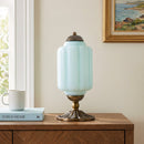 Decorative table lamp with a gold base on a wooden surface, next to a cup and book, with a painting in the background.