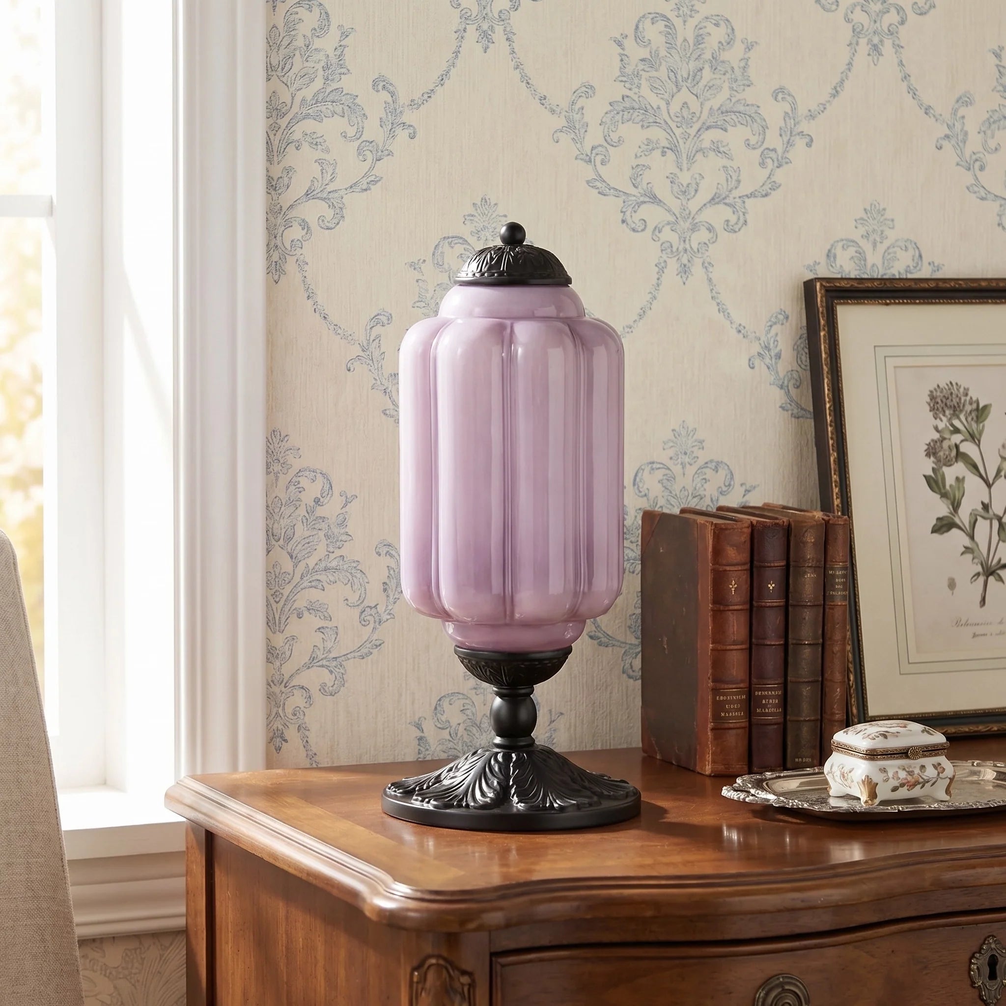 Pink glass lamp on a wooden side table with decorative wallpaper and books in the background