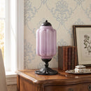 Pink glass lamp on a wooden side table with decorative wallpaper and books in the background