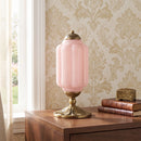 Pink table lamp with gold base on a wooden surface against a decorative wall.
