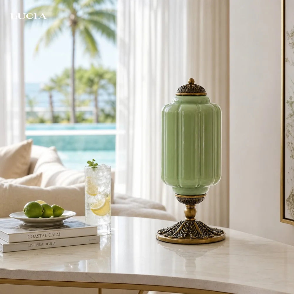 Decorative green lamp with gold base on a table by a window with a view of palm trees and a pool.