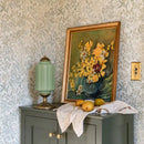 Framed floral painting on a cabinet with a lamp and fruit against a patterned wall.