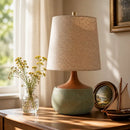 Table lamp with a textured beige shade on a wooden surface, next to a vase with flowers and a small model ship.
