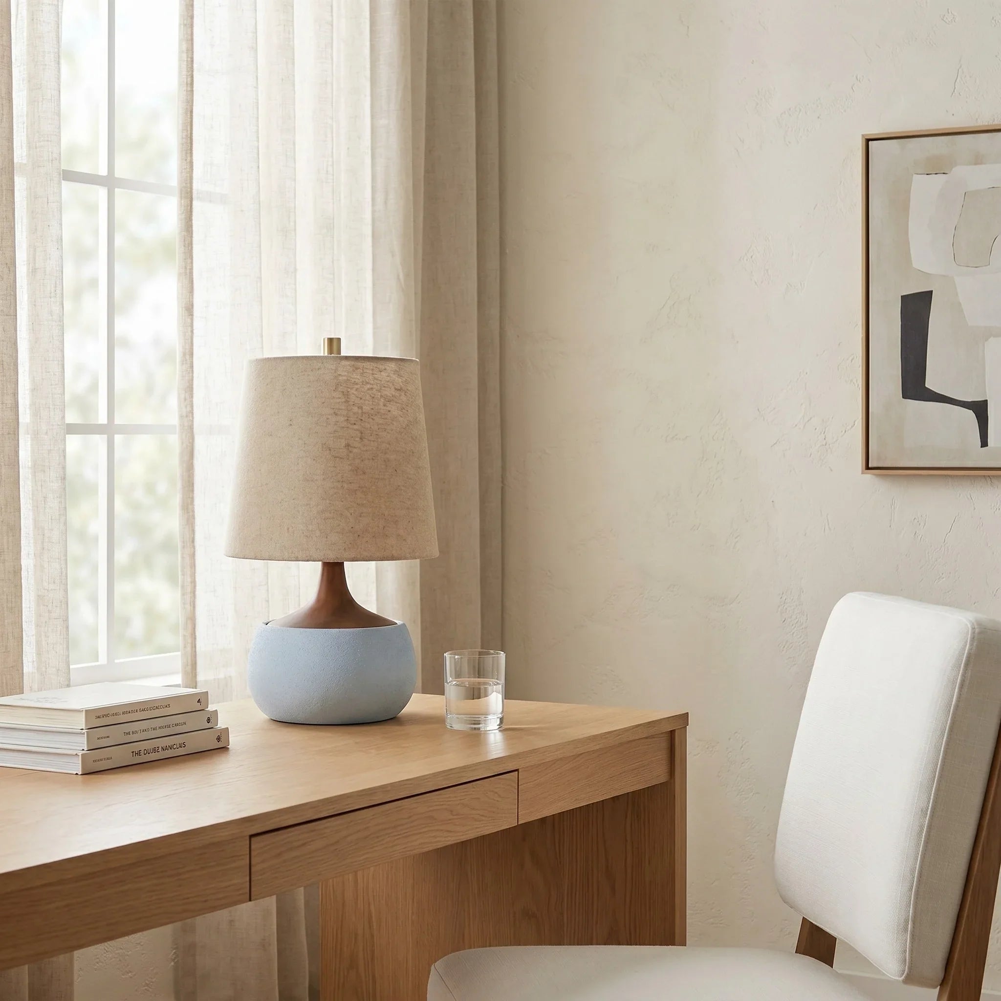 Wooden desk with a lamp and books, white chair, and abstract art on beige wall.