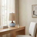 Wooden desk with a lamp and books, white chair, and abstract art on beige wall.