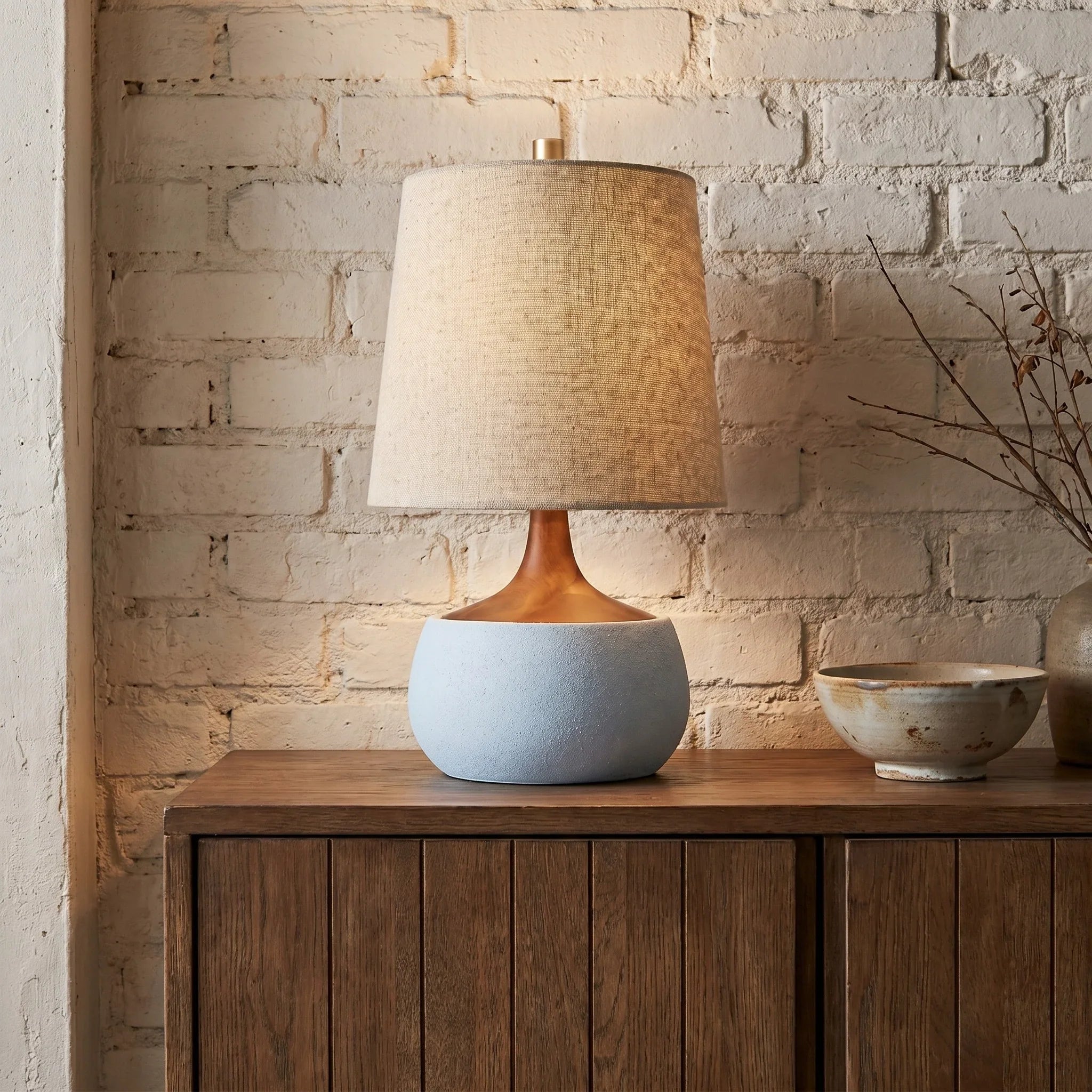 Table lamp with a textured shade on a wooden cabinet against a brick wall.