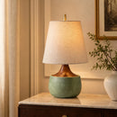 Table lamp with a green base and beige shade on a marble surface, with a plant and framed picture in the background.