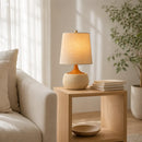 Light wooden side table with a lamp and books next to a beige sofa in a bright room.