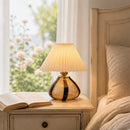Table lamp on a nightstand next to an open book with a window and floral view in the background