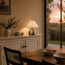 Dining room with a lamp, bowls, and a vase on a cabinet, and a view of a sunset through a window.