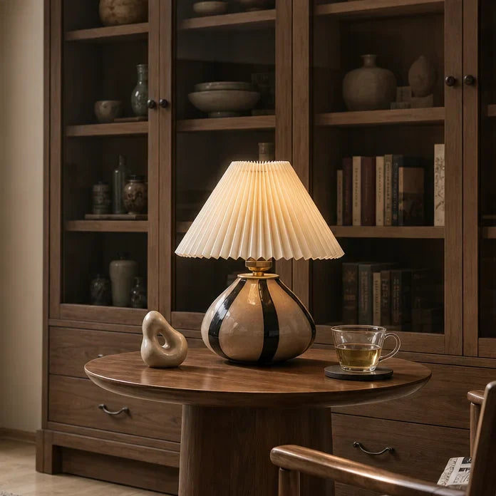 A table lamp with a pleated cream shade and a glossy beige and black ceramic base sits on a round wooden table in a warm, styled room with a bookcase in the background.