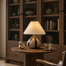 A table lamp with a pleated cream shade and a glossy beige and black ceramic base sits on a round wooden table in a warm, styled room with a bookcase in the background.