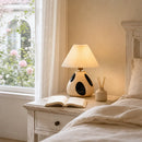 Lamp on a bedside table with an open book and diffuser, next to a window with flowers.