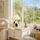 A styled children's bedroom corner with a white side table, pleated table lamp, teddy bear, pillows, and a large window looking out onto a garden.