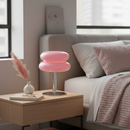 Pink lamp on a wooden nightstand next to a bed with pillows and a blanket.