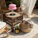 Outdoor setting with wicker side table, pink stool, and basket with towels on a rug.