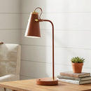 Modern copper desk lamp on a wooden table with a plant and books in the background.