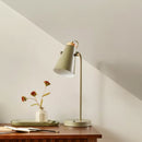 Table lamp with a green shade on a wooden table next to a vase with flowers against a light gray wall.