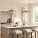 A bright, modern kitchen with two gold pendant lights hanging over a white marble island. The kitchen features white cabinets, a white subway tile backsplash, a gold faucet, and three upholstered bar stools with gold nailhead trim.