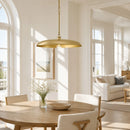 A modern pendant light with a wide, shallow dome shade in brushed gold, hanging over a round wooden dining table in a bright, sunlit dining room.