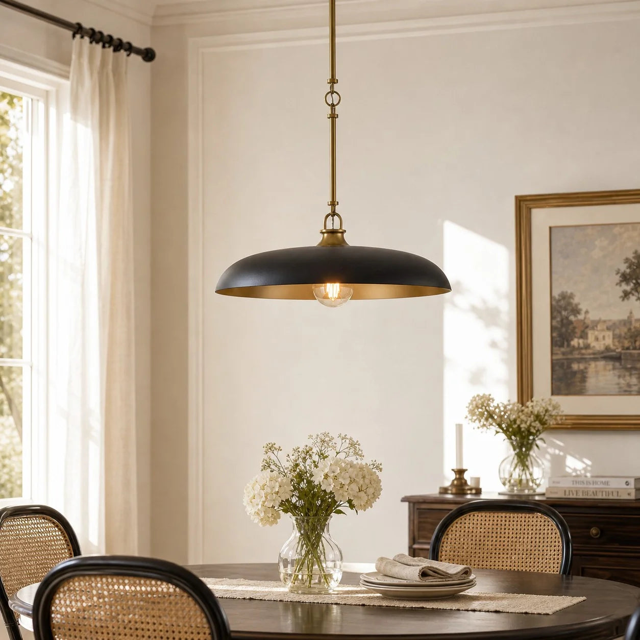 A black and brass pendant light hanging over a dining table in a bright, elegant room with cream walls, woven-back chairs, and floral arrangements.