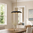 A modern pendant light with a matte black dome-shaped shade and a brushed gold brass rod and trim, hanging over a wooden dining table in a bright, neutral-toned dining room.
