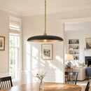A modern pendant light with a matte black dome-shaped shade and a brushed brass suspension chain, hanging over a wooden dining table in a bright, sunlit dining room.