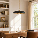 A black and gold pendant light hangs over a wooden desk in a bright, sunlit home office with a bookshelf, window, and leather chair.