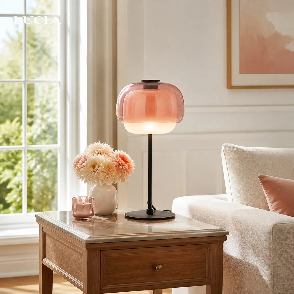 Pink table lamp on a wooden side table with flowers and a glass in a living room.