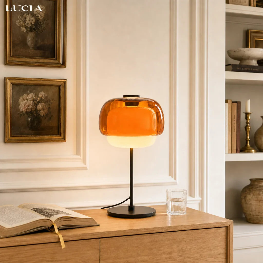 Table lamp with amber glass shade on a wooden surface, with books and decor in the background.