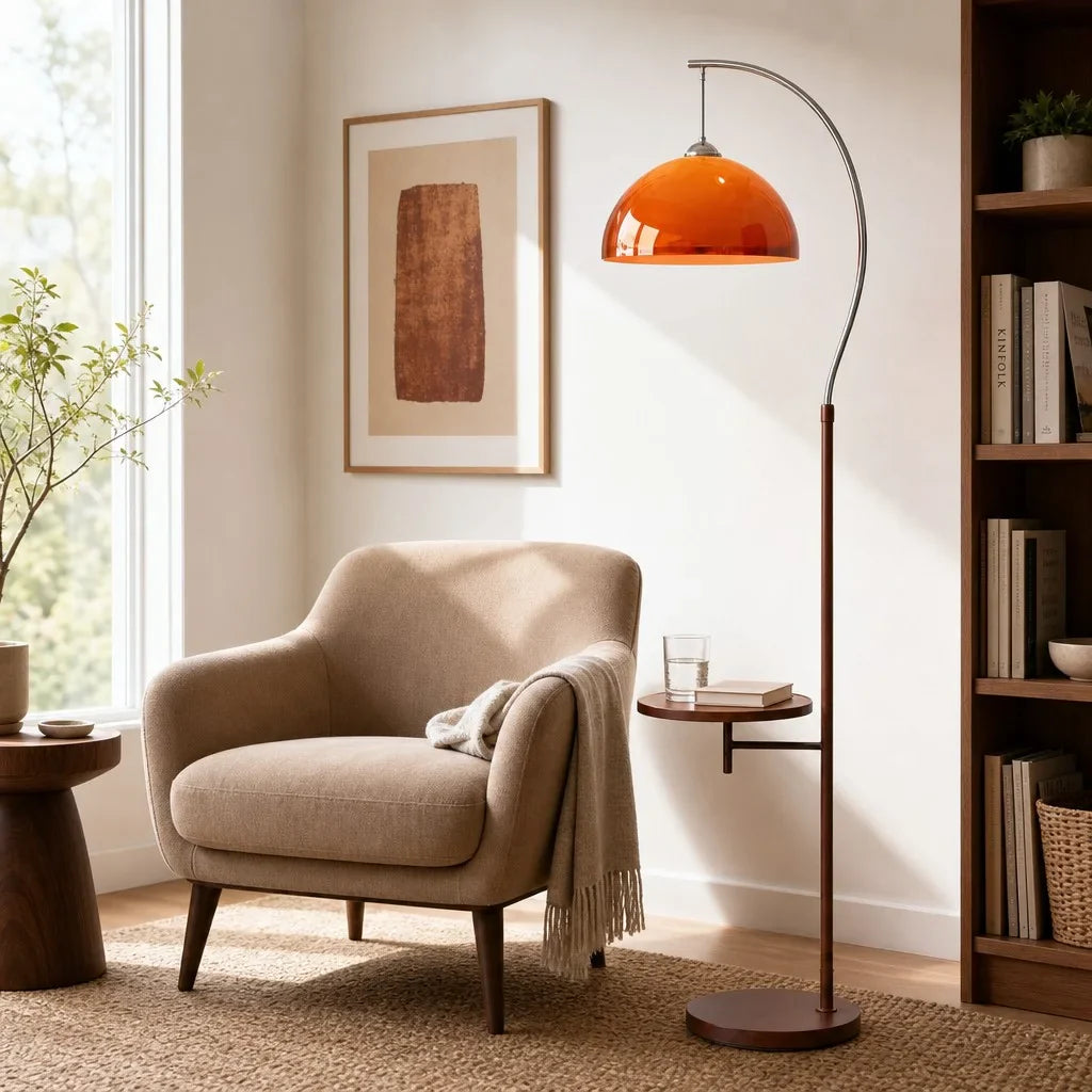 An orange dome floor lamp with a curved dark metal stand and round base, placed beside a beige armchair in a styled living room with a window, framed art, and a bookshelf.
