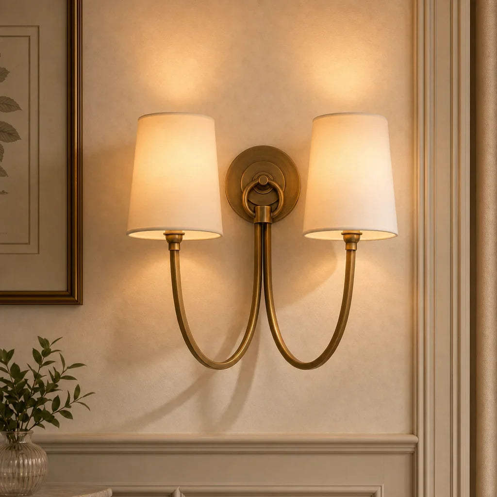 Two-light wall sconce with gold finish and white lampshades on a beige wall.