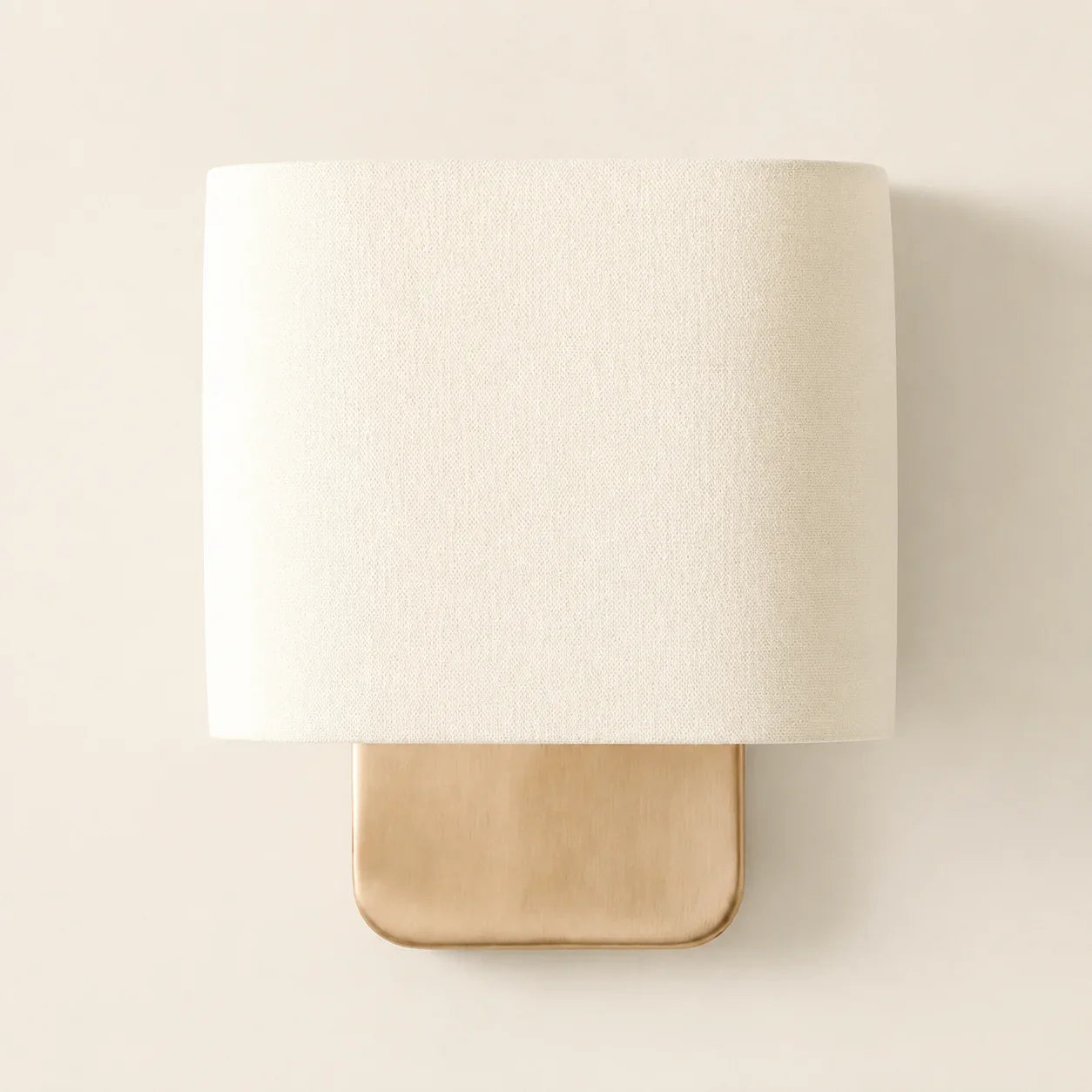 A modern wall sconce light fixture featuring a large, cylindrical off-white fabric shade and a square, brushed gold metal base, shown against a plain beige wall.