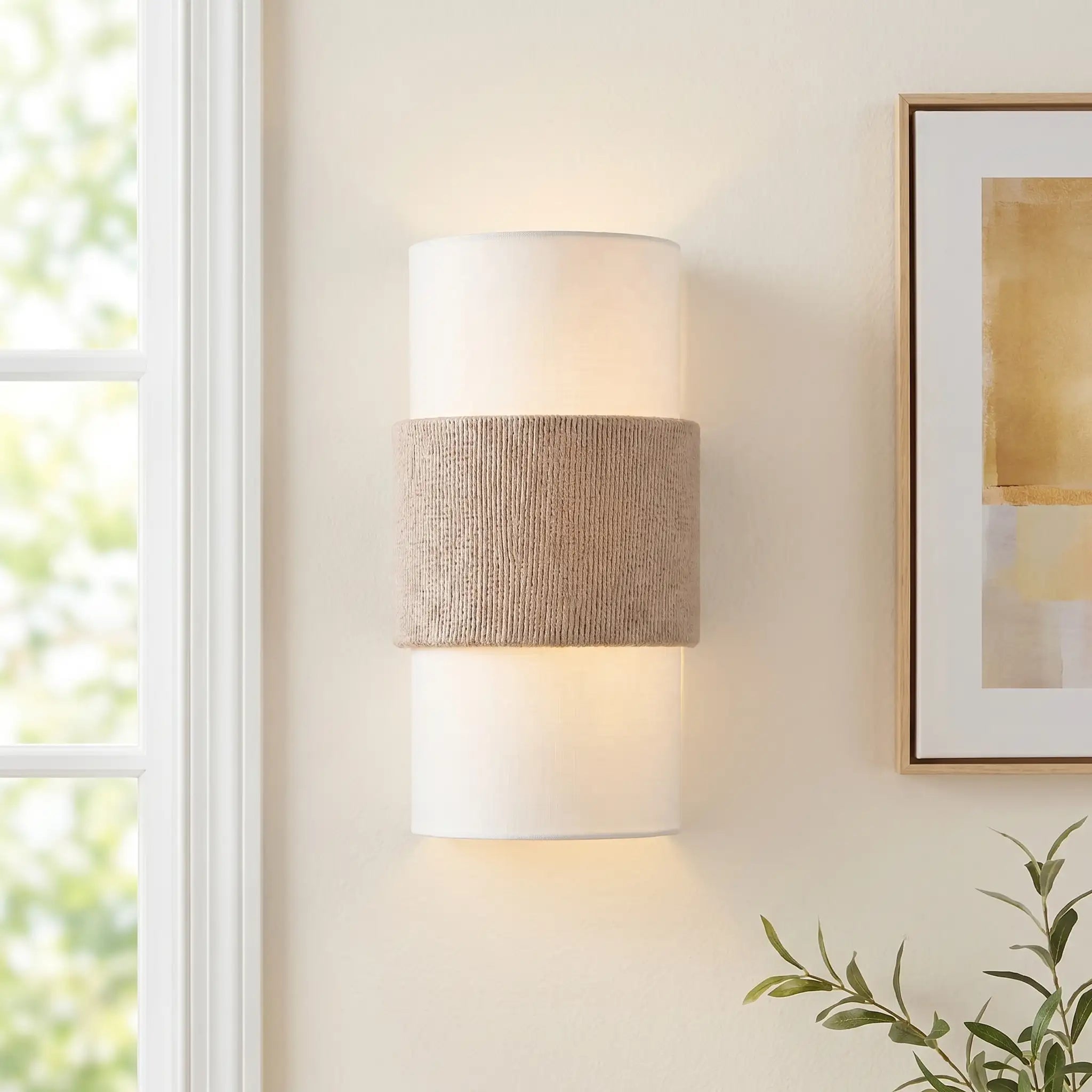 Wall-mounted lamp with textured shade on a wall next to a window and framed picture