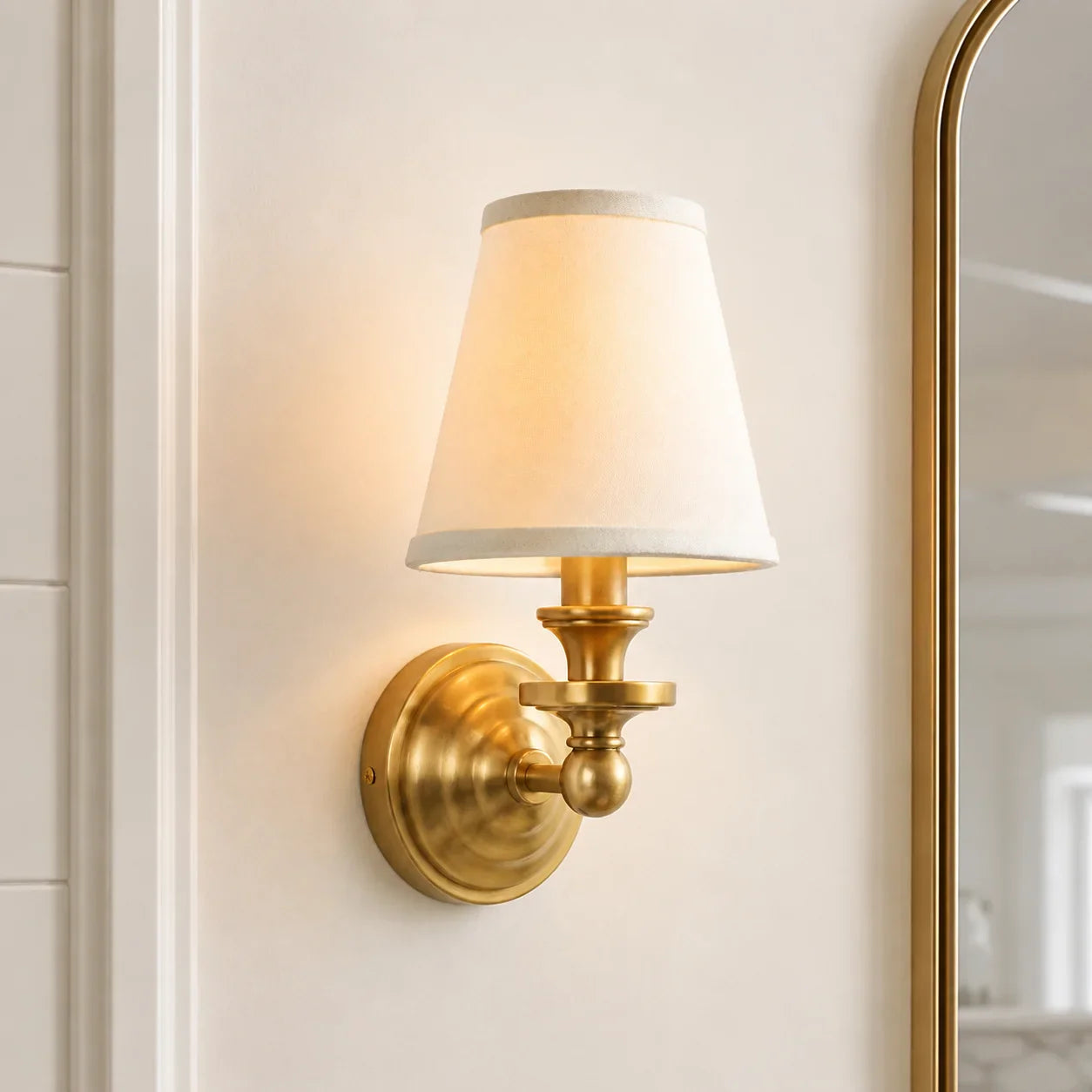 A wall-mounted sconce light fixture with a warm brass/gold metal wall plate and arm, a tapered off-white fabric shade, and a soft warm glow, shown mounted on a cream-colored wall next to a mirror.