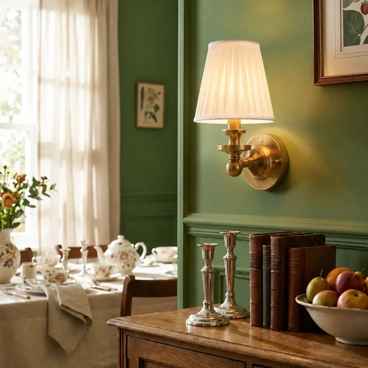 A warm brass wall sconce with a pleated white fabric shade, mounted on a dark green wall above a wooden console table decorated with books, silver candlesticks, and a bowl of fruit.